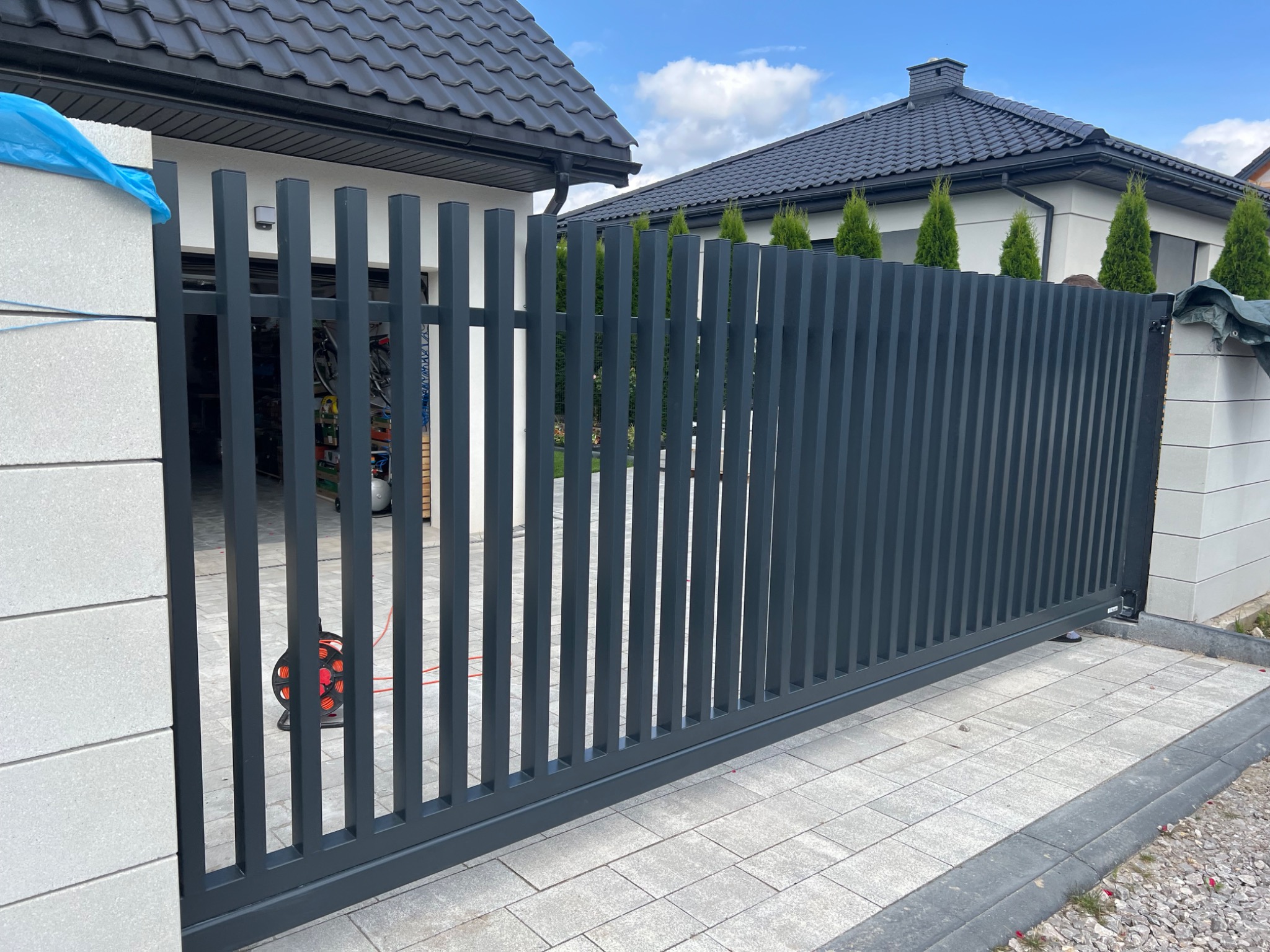 Nowoczesna, przesuwna brama wjazdowa z pionowych profili, szara, na tle domu z ciemnym dachem i tujami. Widoczny fragment kostki brukowej i słupka z bloczków.