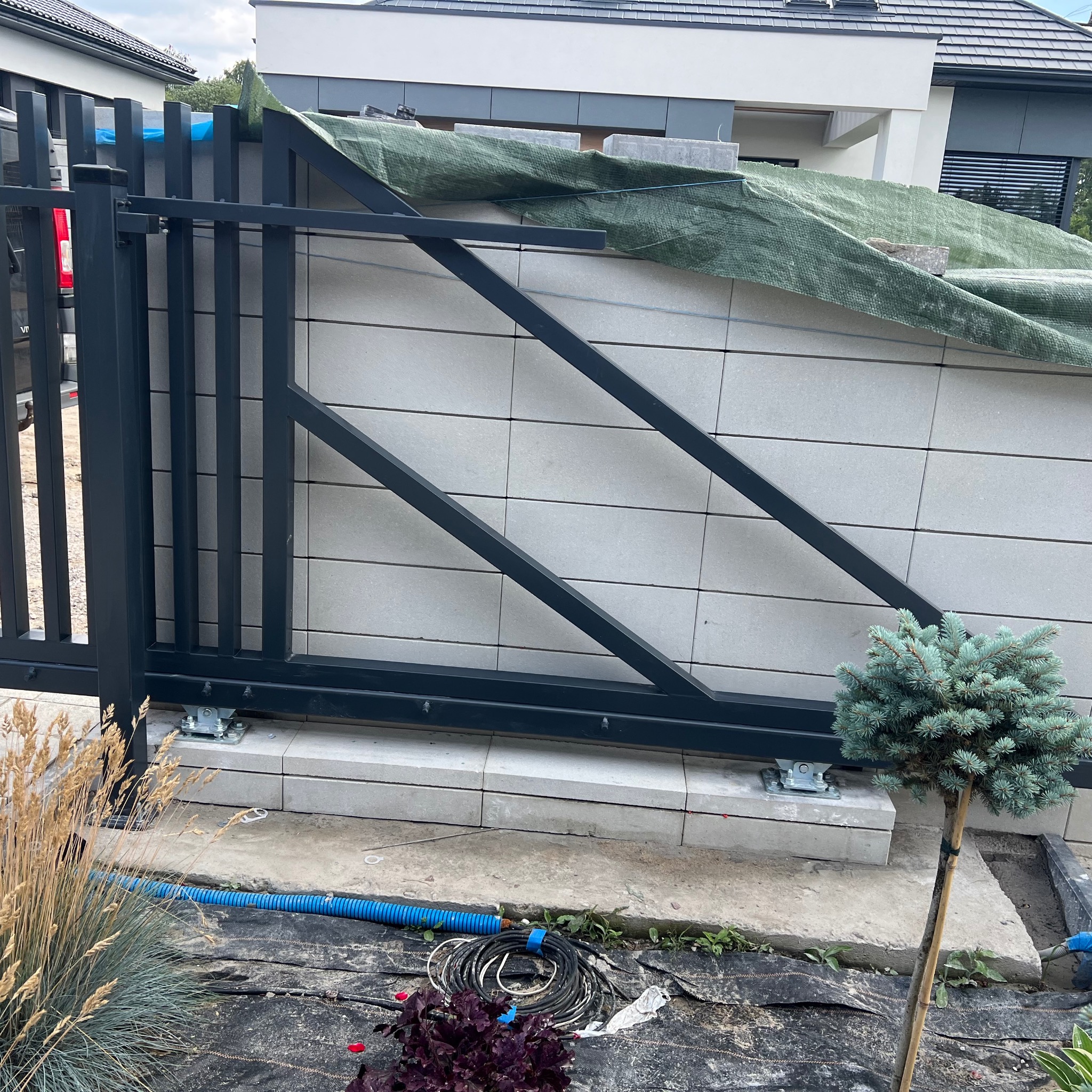 Częściowo zamontowana, przesuwna brama wjazdowa, z widocznym mechanizmem jezdnym. Tło stanowi murowane ogrodzenie z zieloną plandeką na górze.