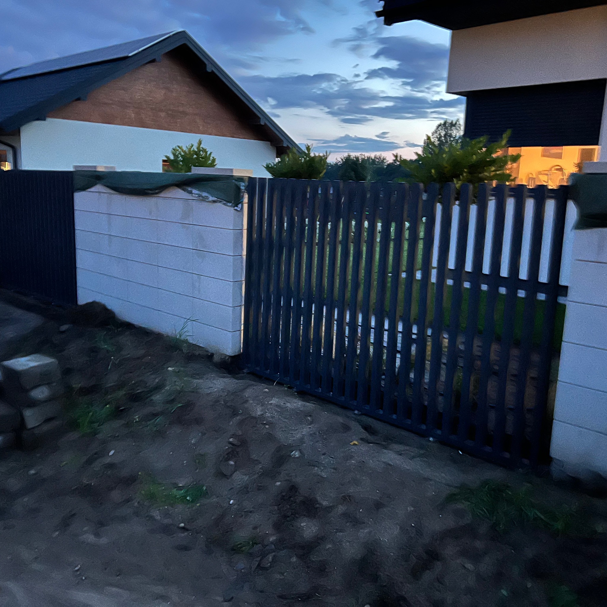 Nowoczesna brama wjazdowa z pionowych, grafitowych sztachet, osadzona na murowanych słupkach. Widok na posesję w wieczornym oświetleniu.