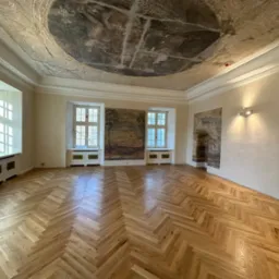 Wnętrze z parkietem w jodełkę, malowidłami na ścianach i suficie. Jasne drewno, kremowe ściany, okna z białymi ramami. Klasyczny, odnowiony styl.