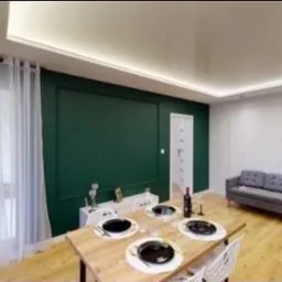 Eleganckie wnętrze po remoncie: butelkowa zieleń ściany, drewniany stół z zastawą, szara sofa. Jasne, przestronne pomieszczenie z nowoczesnym oświetleniem i białymi panelami.