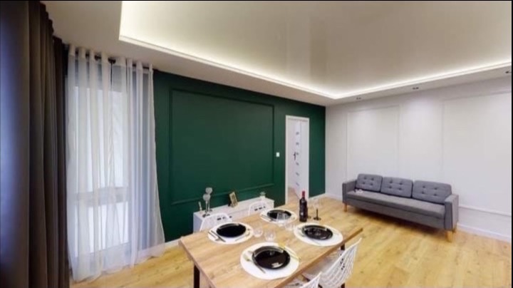 Eleganckie wnętrze po remoncie: butelkowa zieleń ściany, drewniany stół z zastawą, szara sofa. Jasne, przestronne pomieszczenie z nowoczesnym oświetleniem i białymi panelami.