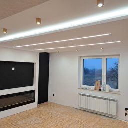 F.H.U TOBMAL TOBIASZ LASOŃ - Nowoczesne wnętrze z oświetleniem LED w suficie podwieszanym, kominkiem elektrycznym i parkietem w jodełkę. Widoczne okno i grzejnik.