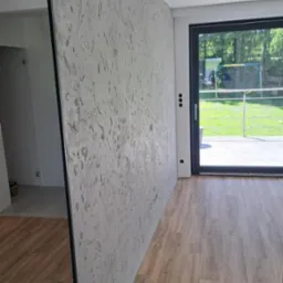 Ściana z dekoracyjną szpachlą w odcieniach szarości, widoczna z perspektywy podłogi z paneli, z oknem balkonowym w tle i fragmentem lustra po lewej.