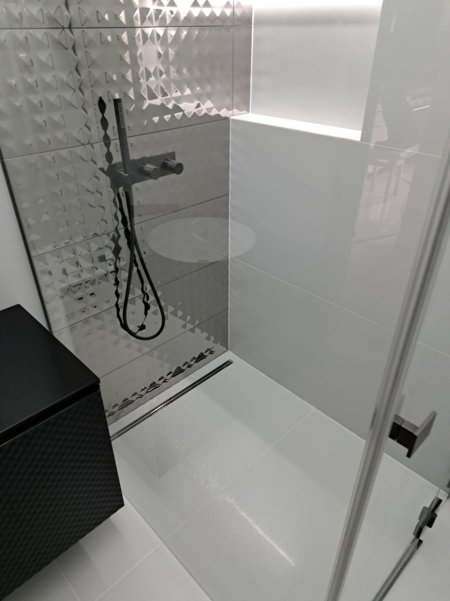 Nowoczesna kabina prysznicowa z czarną armaturą i wzorzystymi szarymi płytkami. Minimalistyczny design, białe płytki podłogowe i częściowo widoczna szafka.