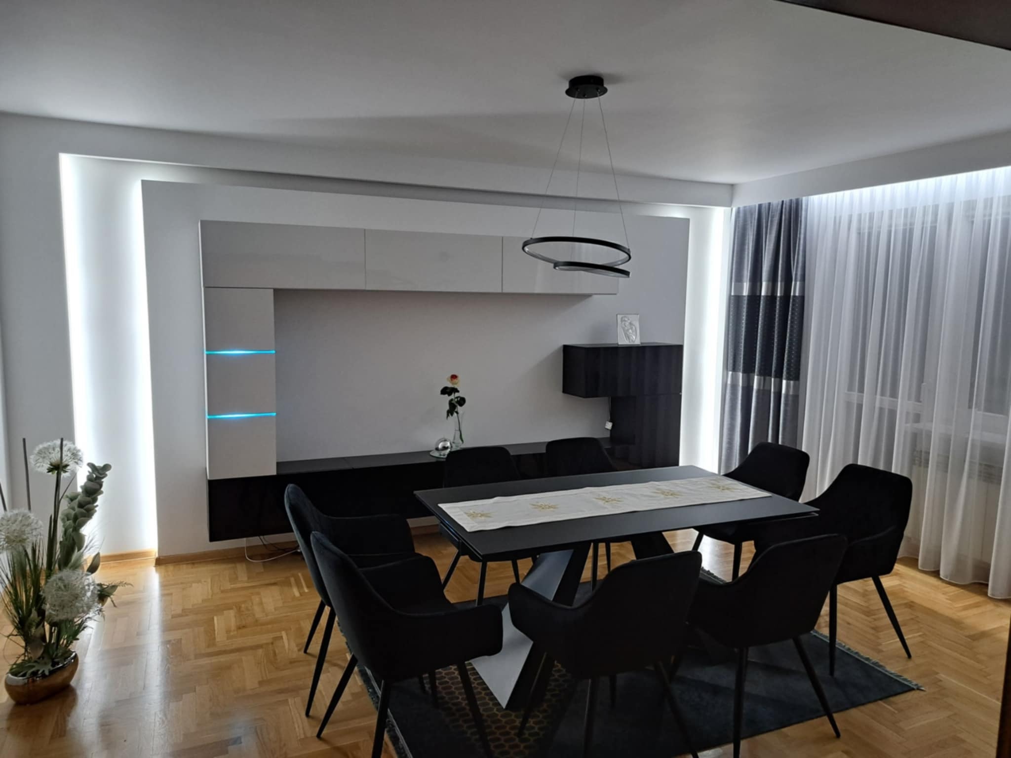 Nowoczesna jadalnia: czarny stół z krzesłami na dywanie, designerska lampa, ściana z podświetleniem LED i dekoracje kwiatowe na parkiecie.