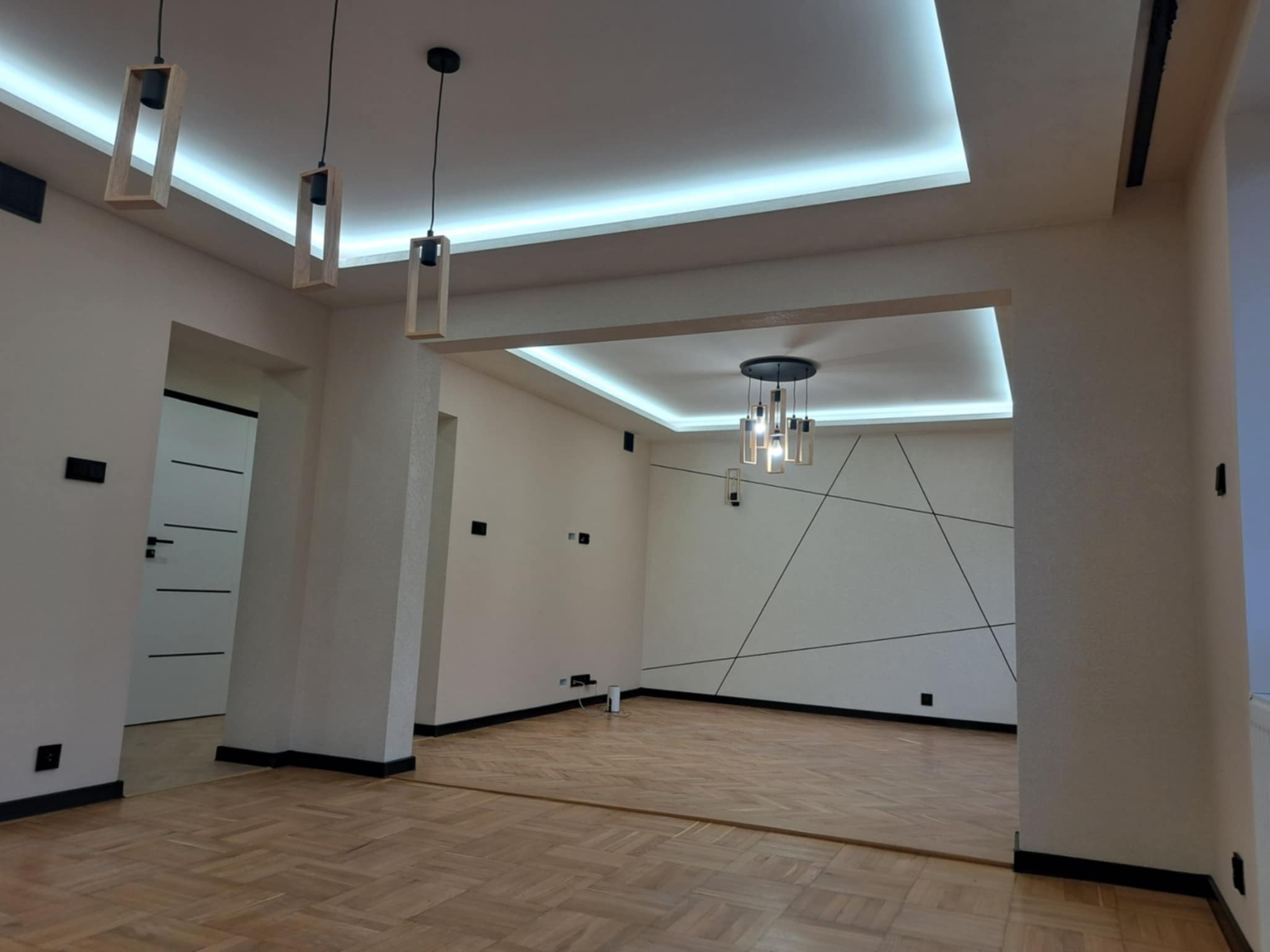 Nowoczesne wykończenie wnętrza: parkiet w jodełkę, oświetlenie LED w suficie podwieszanym i minimalistyczne lampy wiszące w wyremontowanym salonie.