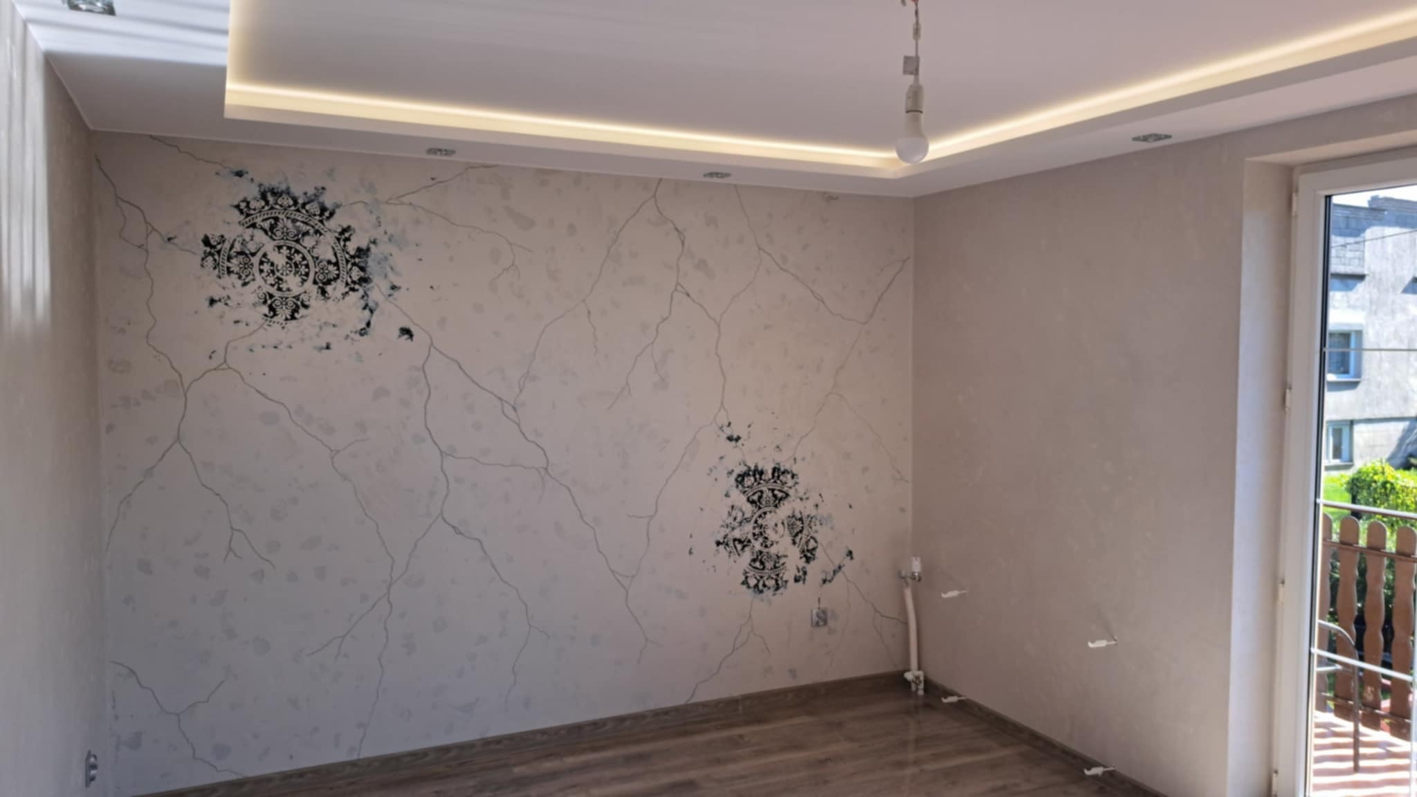 Wykończone wnętrze pokoju z dekoracyjną ścianą w stylu crackle z ornamentami, drewnianą podłogą i oświetleniem LED podsufitowym. Widok na balkon i budynek z okna.