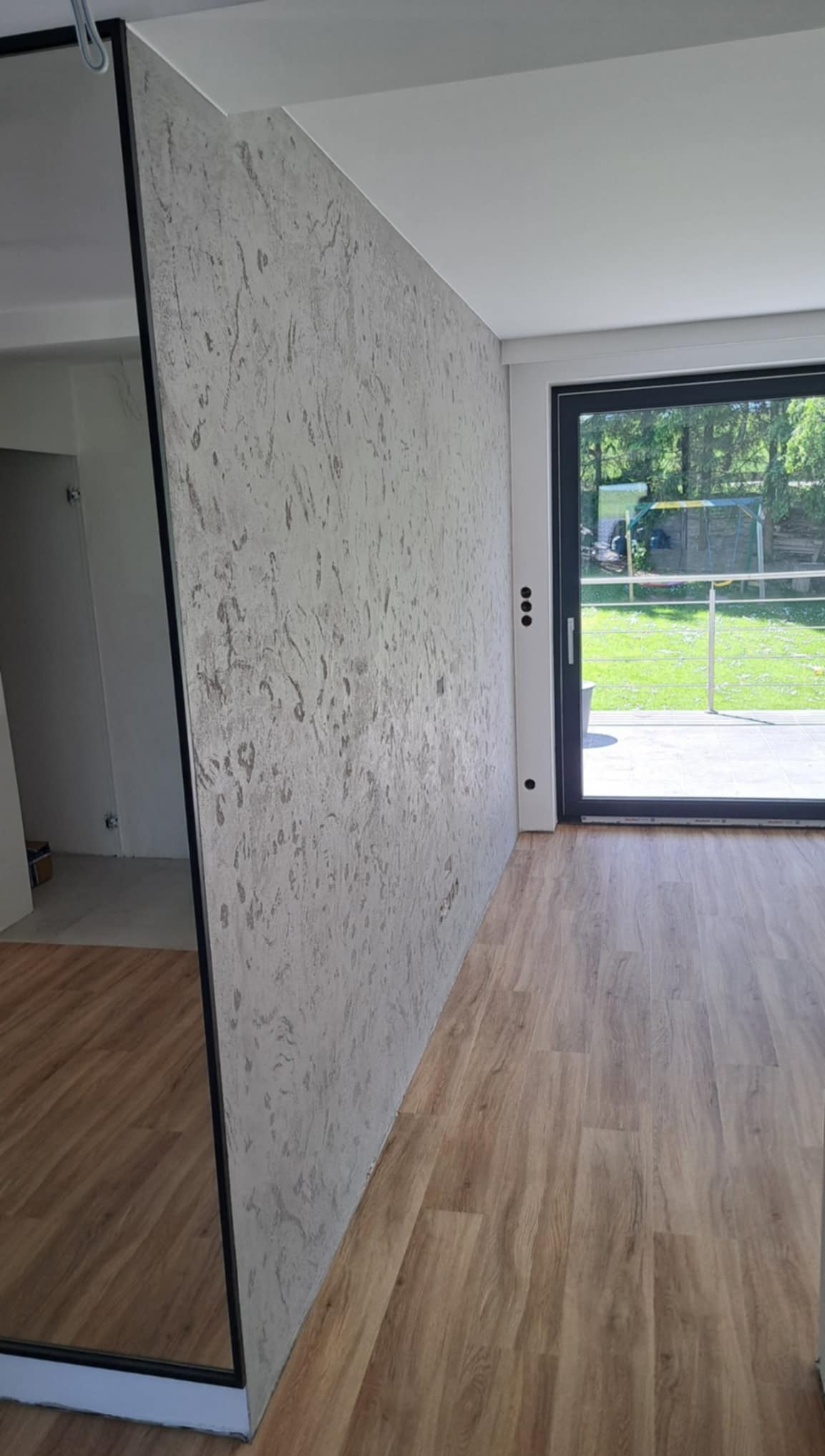 Ściana z dekoracyjną szpachlą w odcieniach szarości, widoczna z perspektywy podłogi z paneli, z oknem balkonowym w tle i fragmentem lustra po lewej.