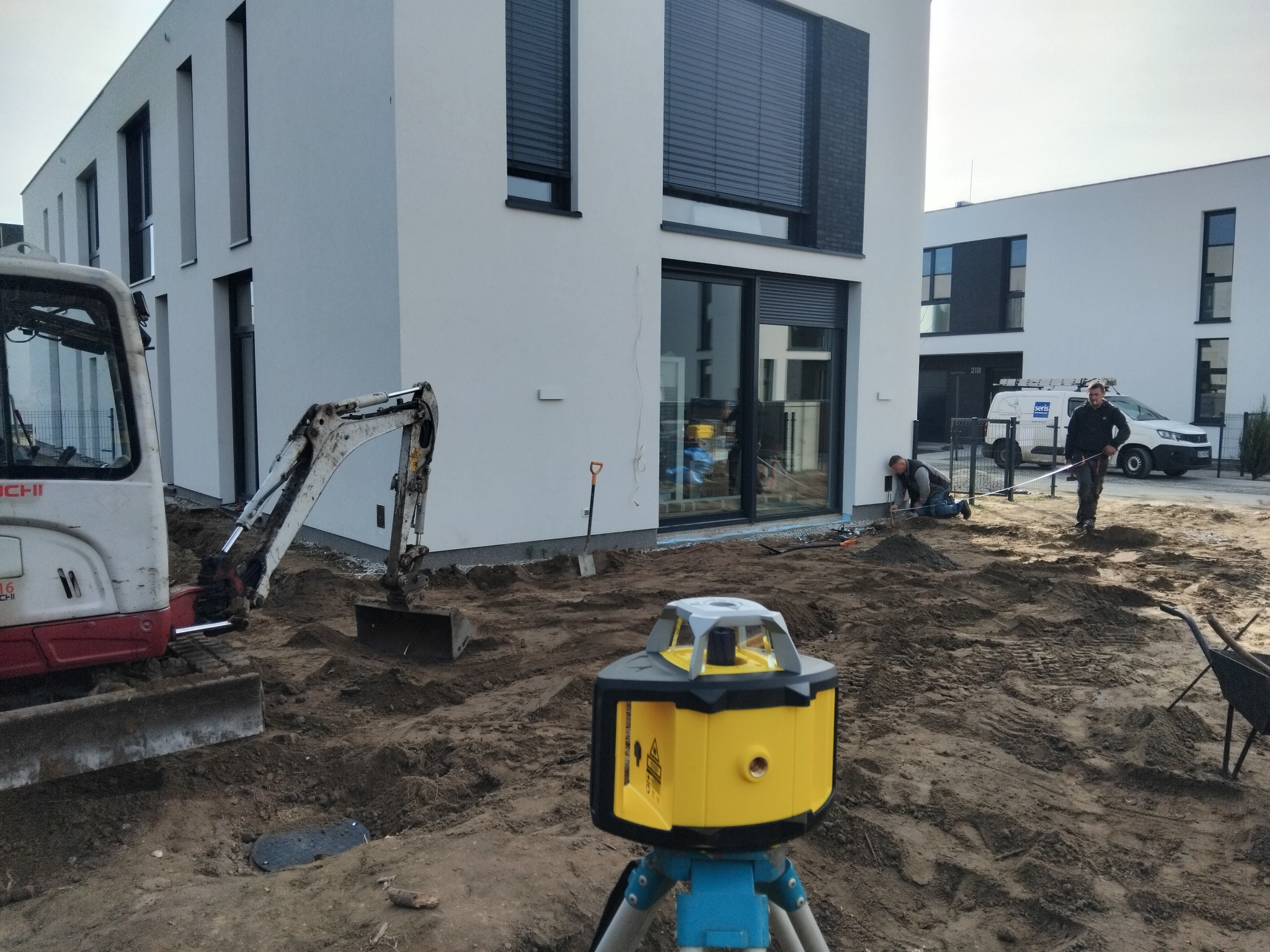 Prace ziemne przy nowoczesnym budynku: mini koparka, niwelator laserowy na statywie, pracownicy wyrównujący teren. W tle biała zabudowa szeregowa i samochód dostawczy.
