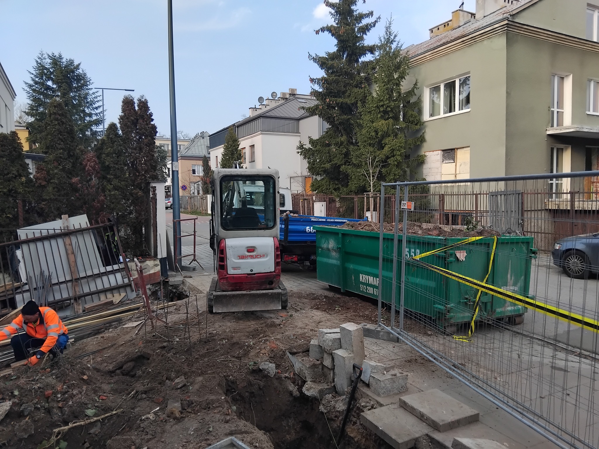 Prace ziemne w toku: robotnik w pomarańczowej kurtce przy wykopie, obok minikoparka Takeuchi TB216, kontener Krymap i wygrodzenie z metalowej siatki.