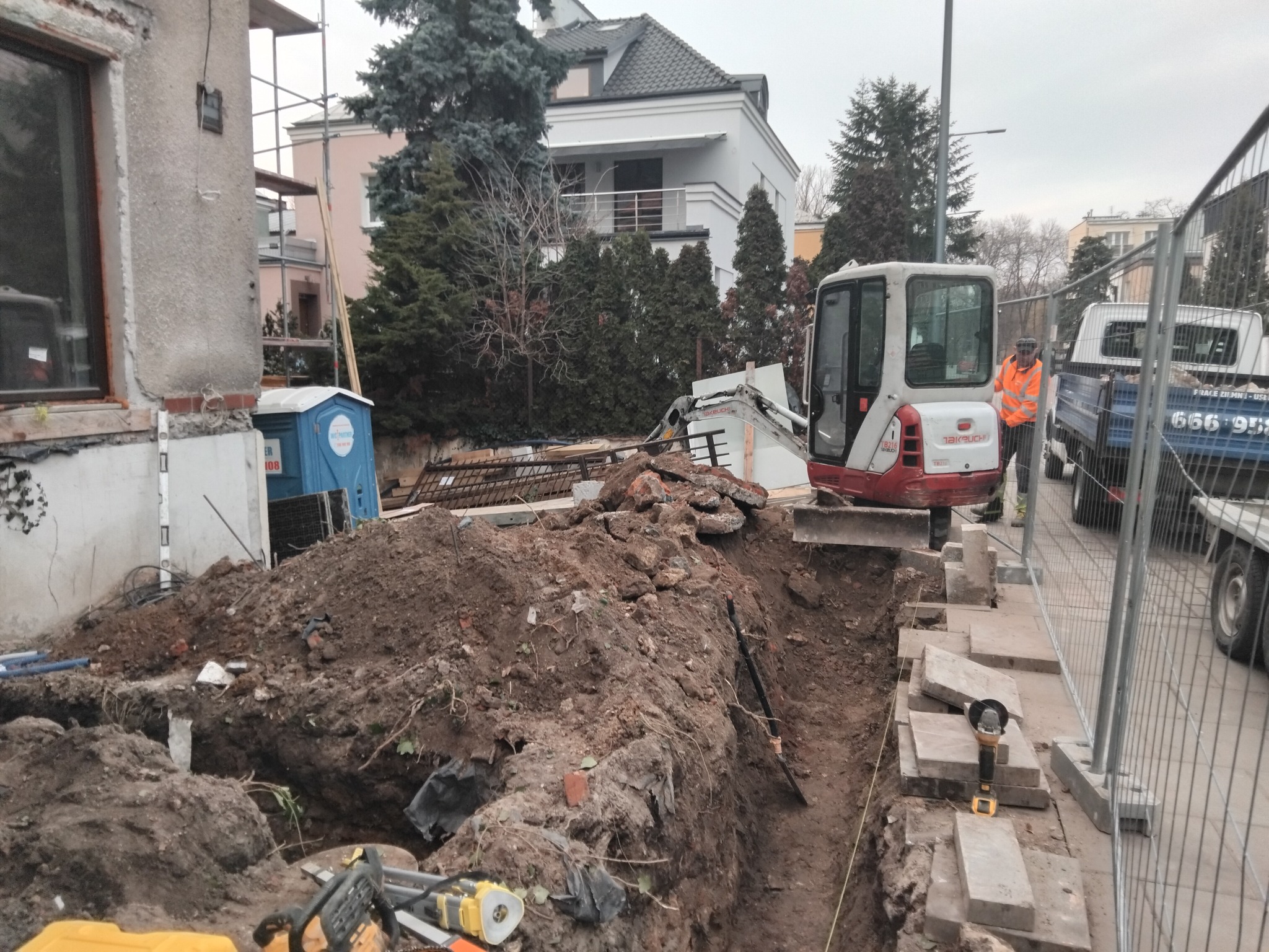 Wykop pod fundamenty z mini koparką Takeuchi, widoczny pracownik w pomarańczowej kamizelce. Na pierwszym planie narzędzia i wykopana ziemia.