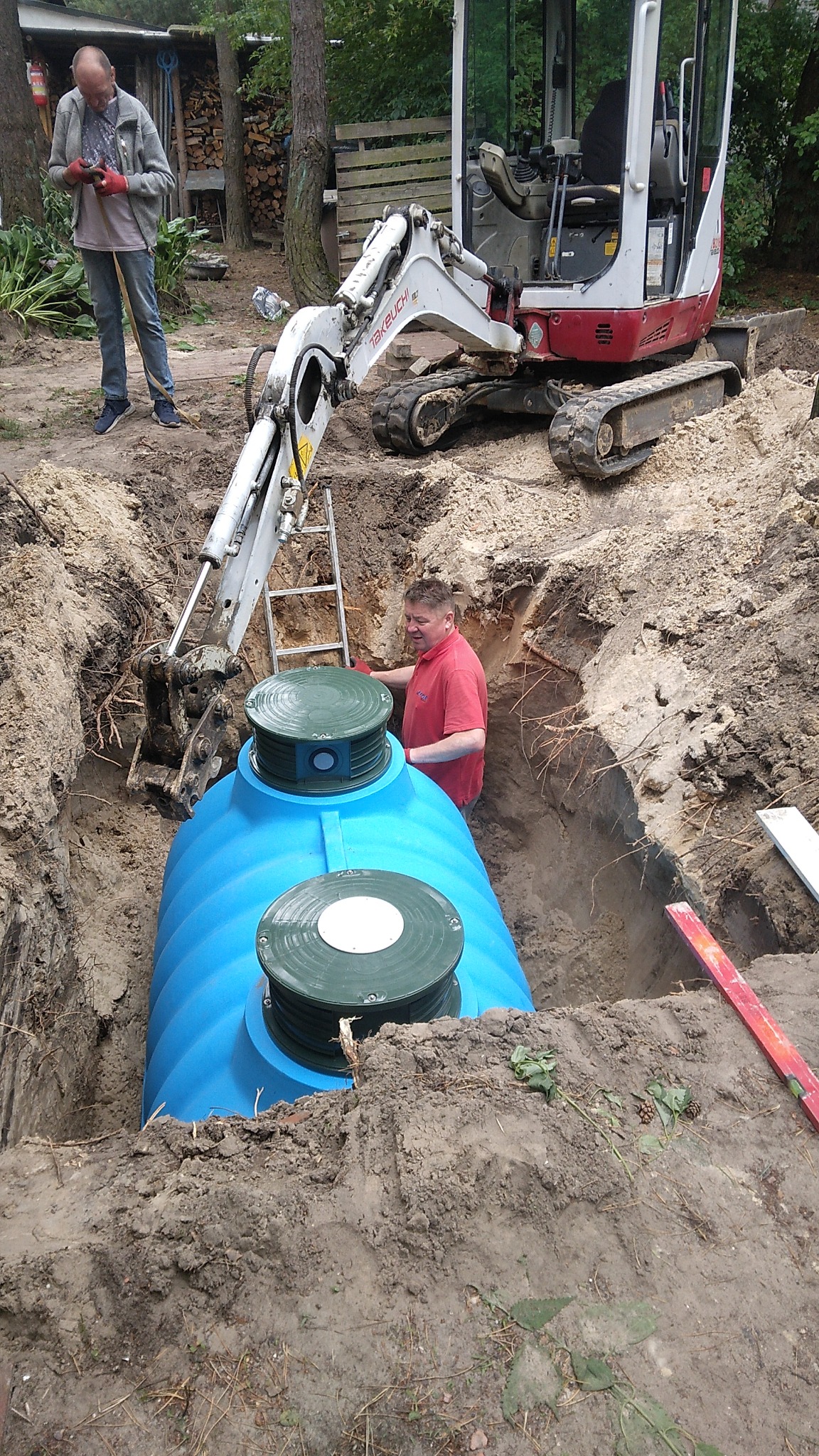 Montaż niebieskiego zbiornika na szambo w wykopie, z użyciem minikoparki Takeuchi. Pracownik w wykopie, drugi przy koparce. Teren zadrzewiony, poziomica na ziemi.