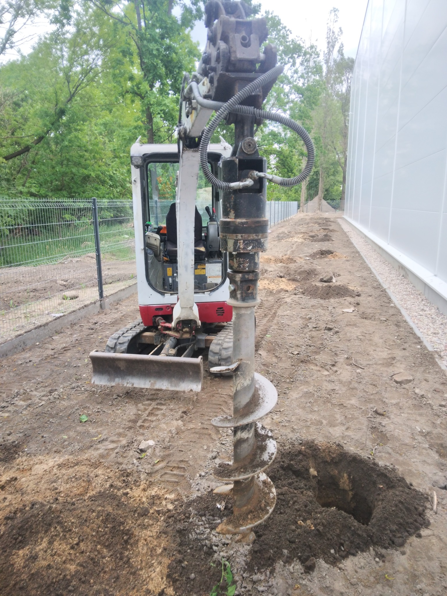 Mini koparka z wiertnicą świdrową wykonuje serię otworów w ziemi wzdłuż ogrodzenia i ściany budynku. Widoczny krajobraz z drzewami w tle.