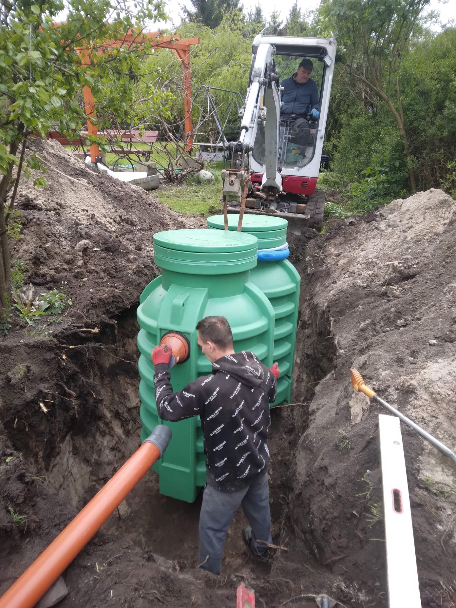 Montaż zielonego zbiornika na szambo w wykopie. Pracownik łączy rurę, a w tle minikoparka. Widoczne narzędzia i poziomica.