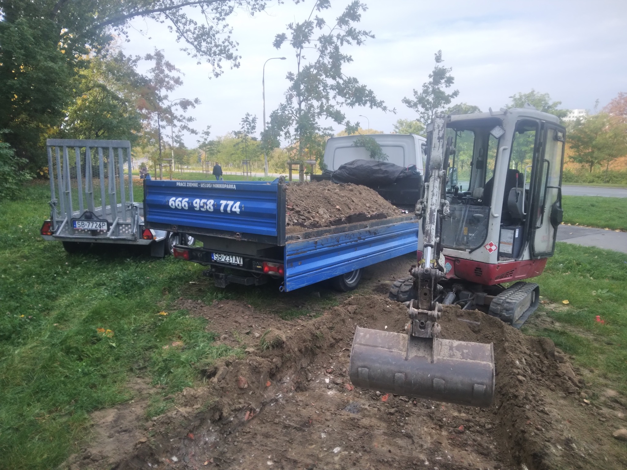 Mini koparka z łyżką, wykopany rów, przyczepka i auto dostawcze z ziemią. Usługi minikoparką. Telefon kontaktowy na burcie przyczepy. Jesienna sceneria.