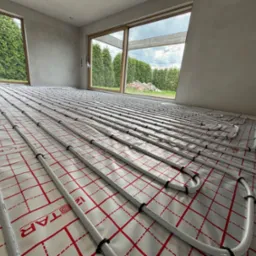 Białe rury ogrzewania podłogowego równolegle ułożone na folii z czerwoną kratą, widok z góry, surowy stan wykończenia wnętrza z oknami na zieleń.