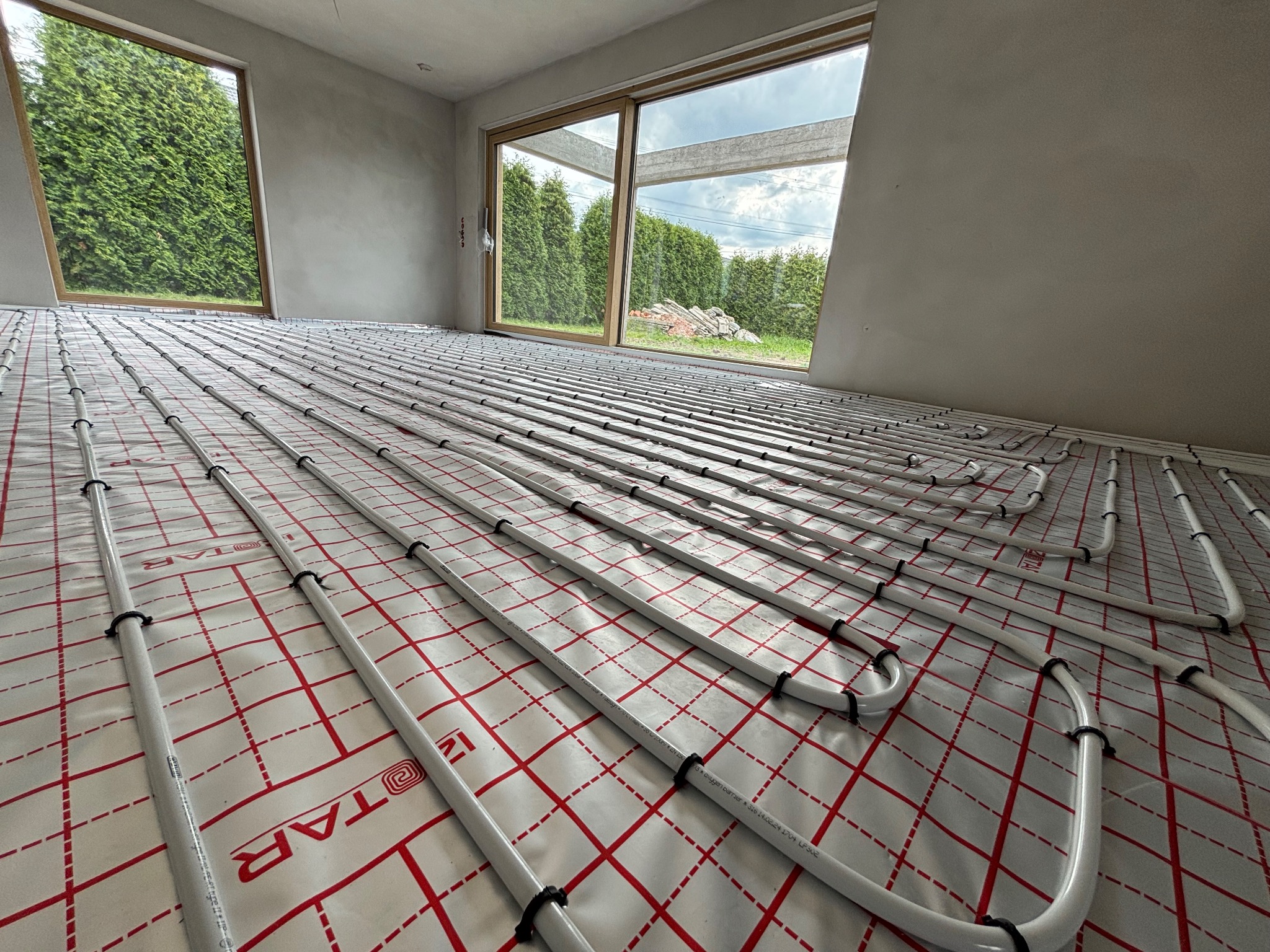 Białe rury ogrzewania podłogowego równolegle ułożone na folii z czerwoną kratą, widok z góry, surowy stan wykończenia wnętrza z oknami na zieleń.
