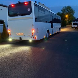 ŁACH ADRIAN Lider Trans Przewóz Osób Autokarem - Biały autokar Irisbus, widok z tyłu, oświetlony światłami stopu i cofania, zaparkowany na asfalcie. Widoczny numer rejestracyjny i znak ograniczenia prędkości do 100 km/h.