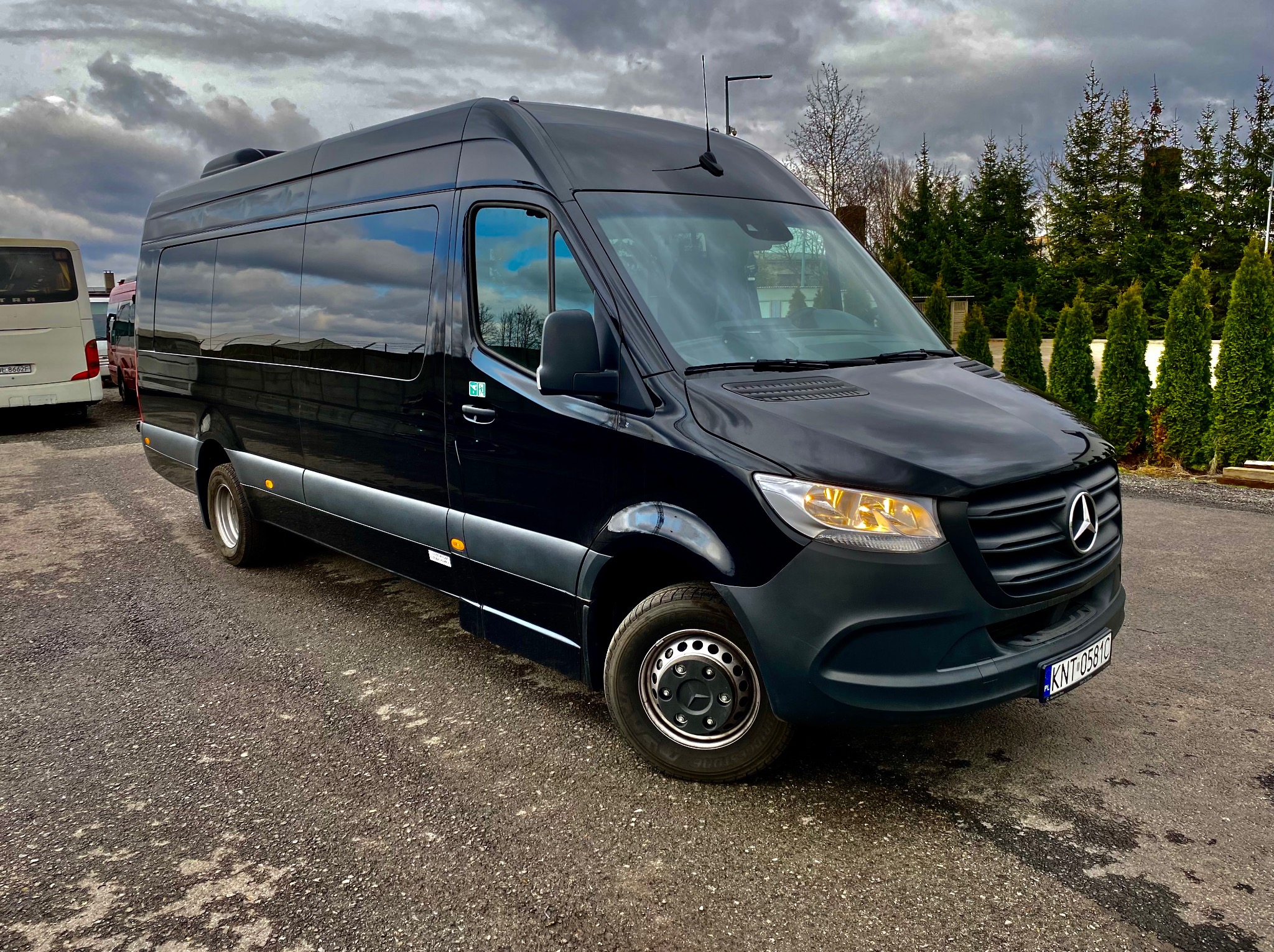 Czarny, lśniący Mercedes Sprinter z anteną na dachu, zaparkowany na asfalcie, z widocznymi detalami jak felgi i tablica rejestracyjna, na tle pochmurnego nieba i zieleni.