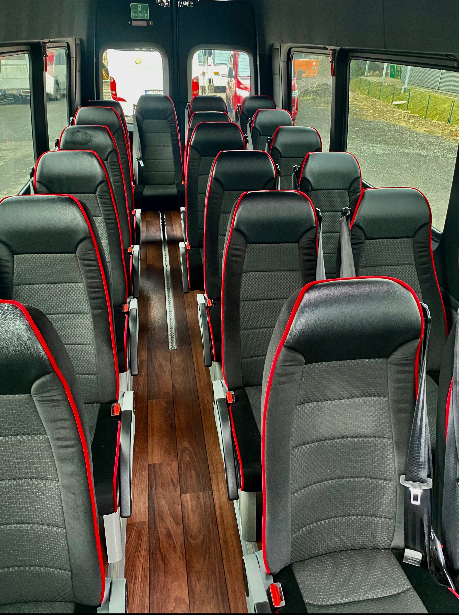 Wnętrze busa pasażerskiego z rzędami foteli obitych czarną skórą z czerwonymi akcentami, widok z perspektywy środka pojazdu, drewnopodobna podłoga.