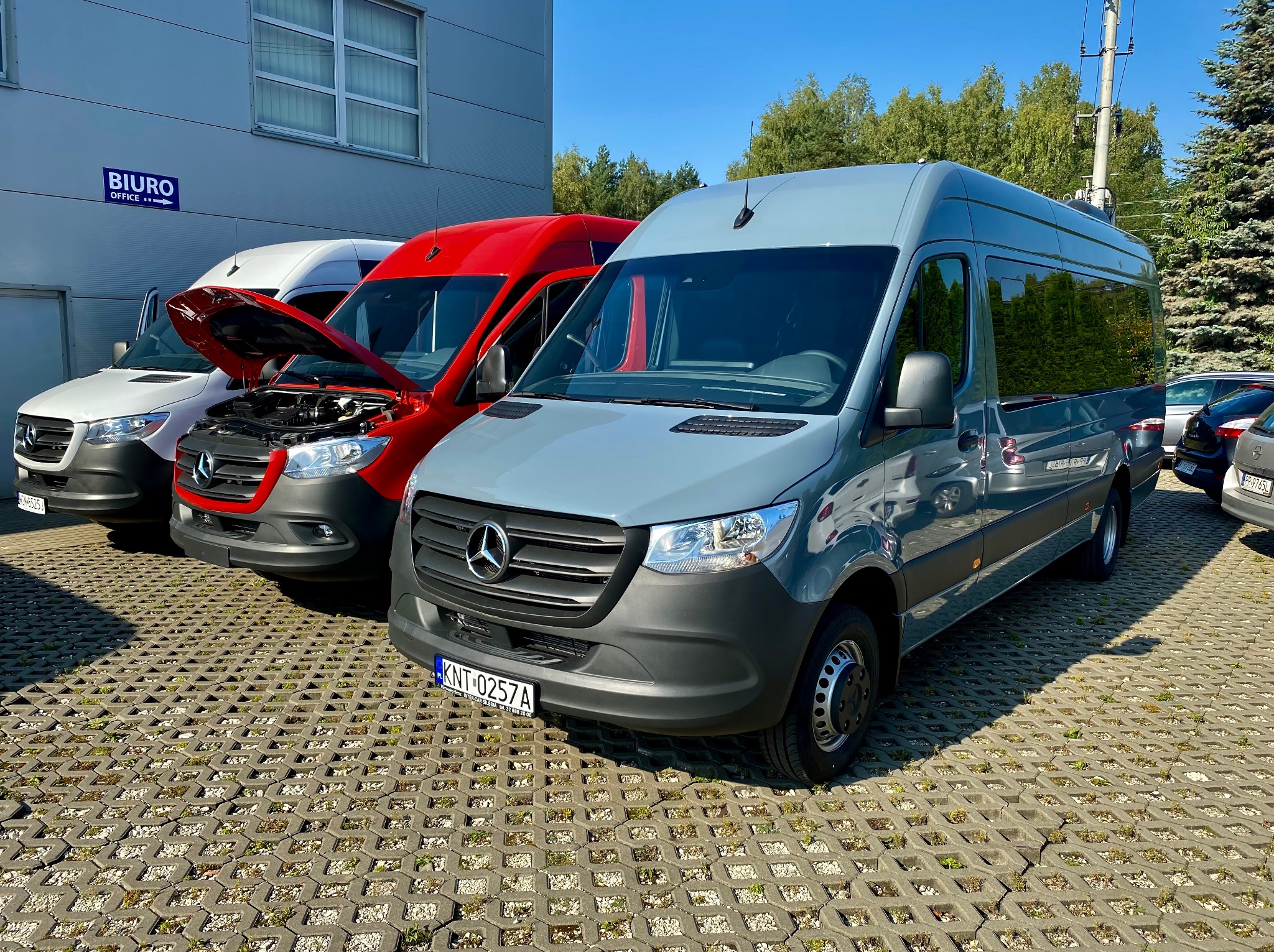 Flota busów Mercedes Sprinter w kolorach białym, czerwonym i szarym, zaparkowana na ażurowym parkingu przed budynkiem biurowym w słoneczny dzień. Jeden z busów ma otwartą maskę.