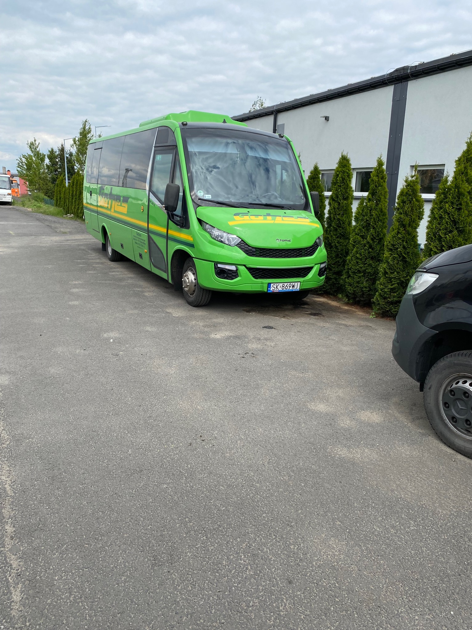 Zielony bus Iveco z żółtymi pasami i napisem, zaparkowany na parkingu obok budynku z tujami. Widoczny fragment czarnego samochodu.