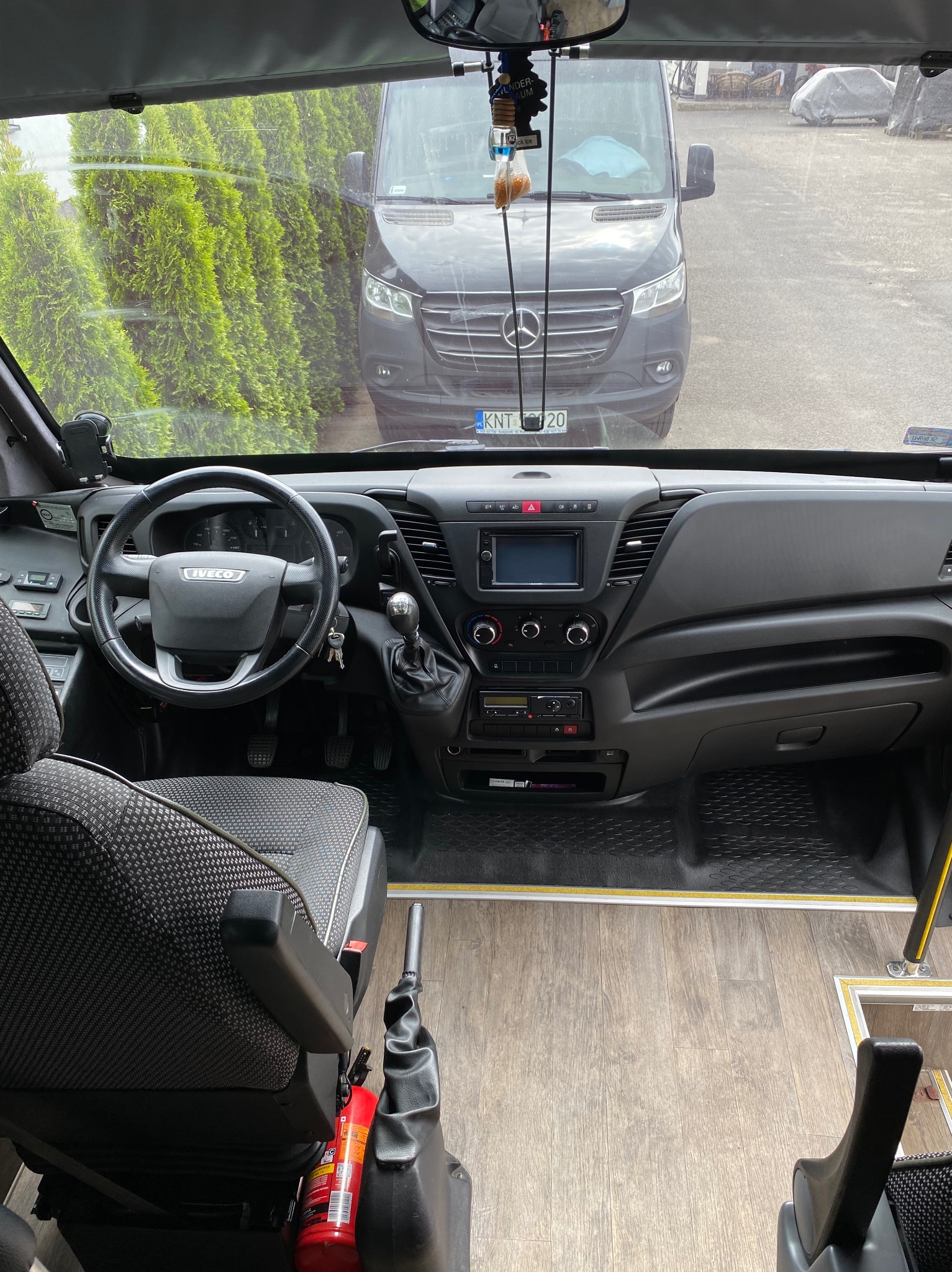 Wnętrze busa pasażerskiego z widokiem na deskę rozdzielczą, fotel kierowcy i szybę przednią, za którą stoi czarny bus marki Mercedes.