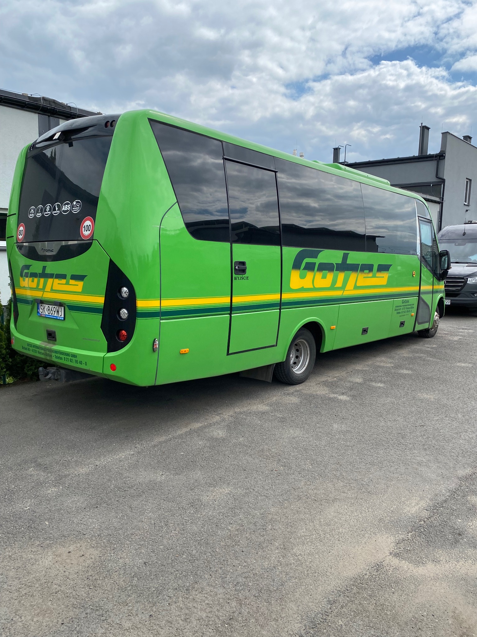 Zielony minibus z żółtymi pasami i napisem 'Gotzes' na tle szarego budynku i pochmurnego nieba. Widoczny fragment parkingu i tylna część pojazdu.