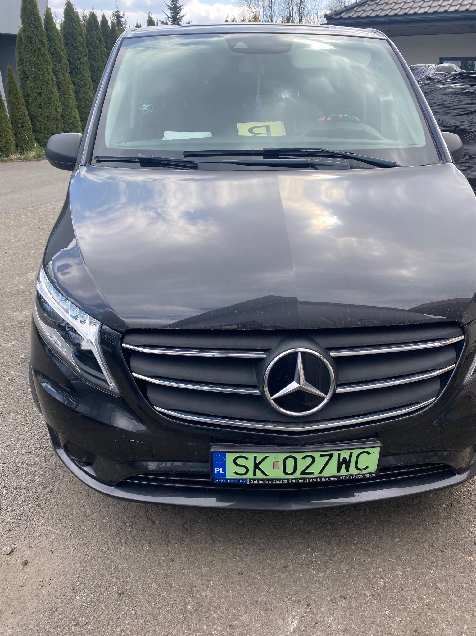 Czarny Mercedes Vito z zieloną tablicą rejestracyjną SK 027WC na tle szarym, z widocznym logo marki i fragmentem ulicy. Na desce rozdzielczej dekoracja z kwiatów.