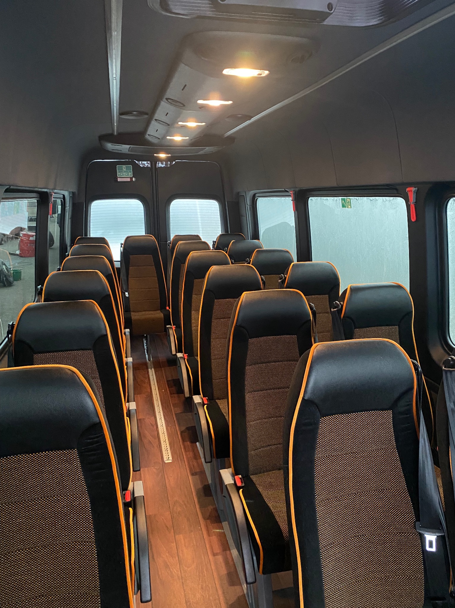 Wnętrze autobusu z rzędami czarno-pomarańczowych foteli, drewnianą podłogą i oknami z widokiem na zewnątrz. Widoczne oświetlenie sufitowe i znaki bezpieczeństwa.