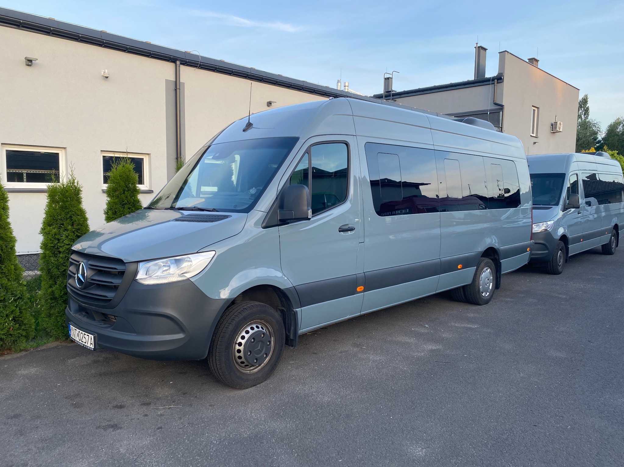 Szary bus marki Mercedes-Benz Sprinter z przyciemnianymi szybami, zaparkowany przed budynkiem. Widoczny fragment drugiego, identycznego pojazdu.