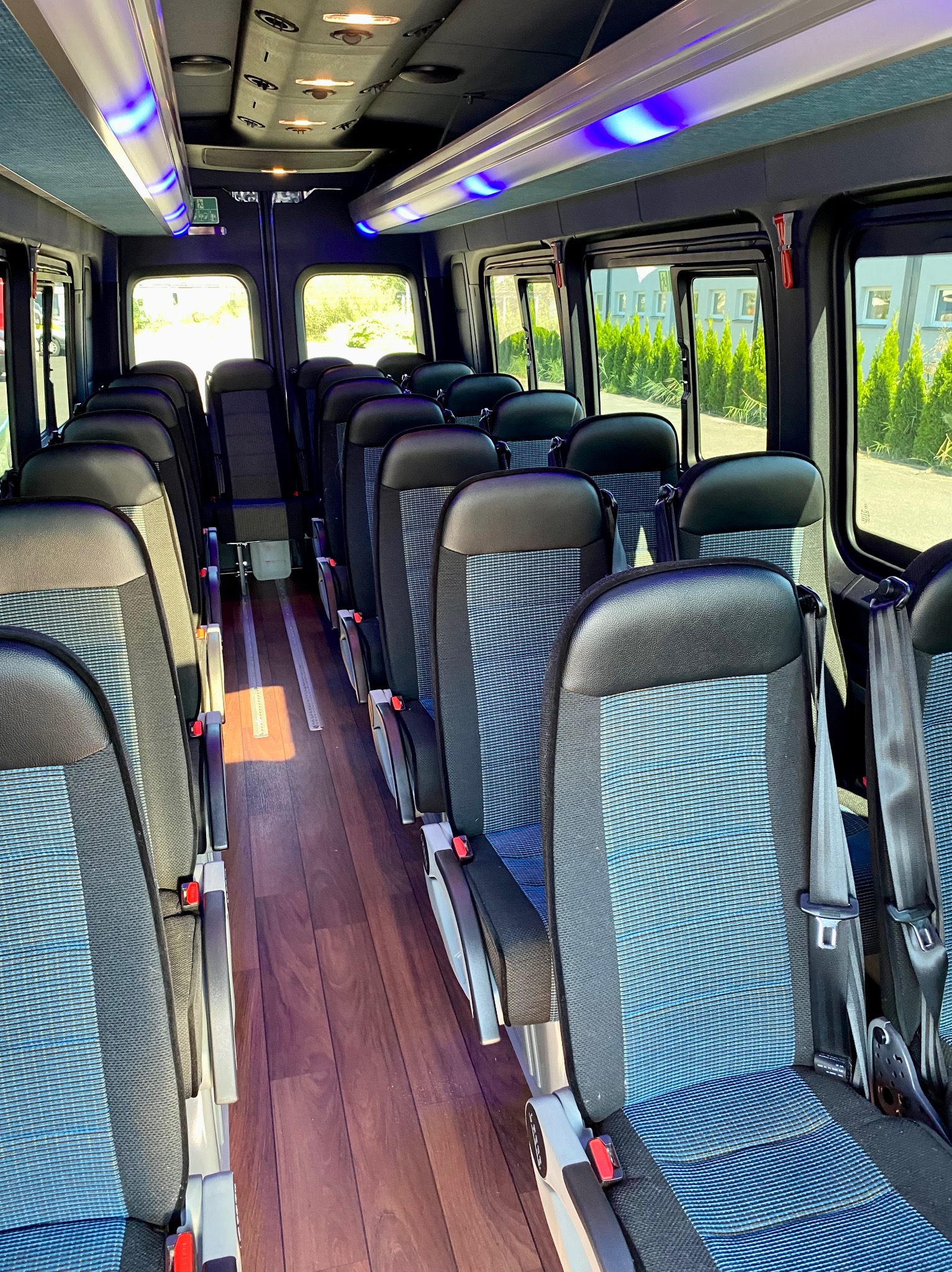 Wnętrze autobusu z rzędami foteli z niebieskimi wstawkami i drewnianą podłogą. Widoczne okna z zielenią na zewnątrz i oświetlenie sufitowe.