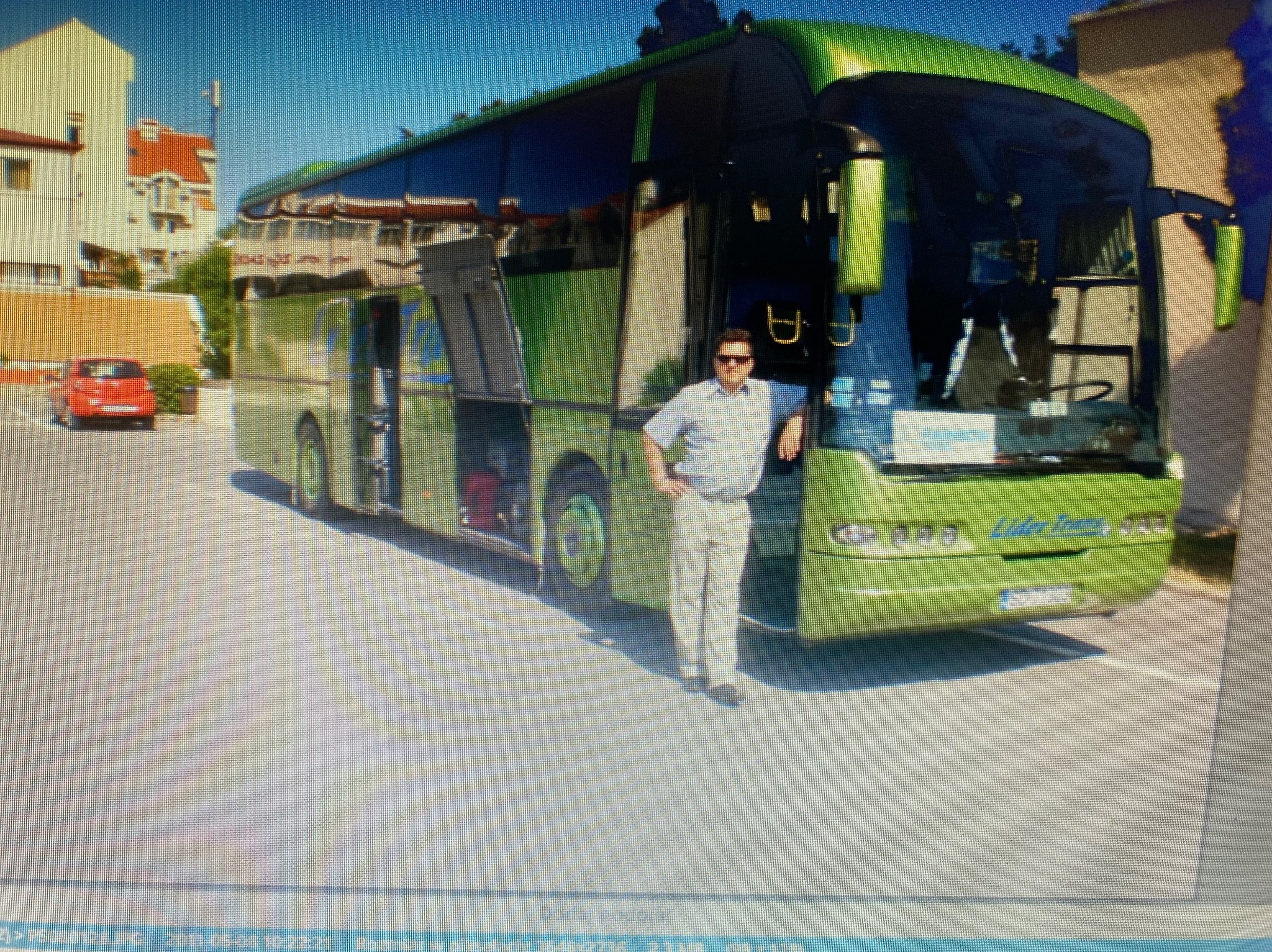 Zielony autokar Lider Trans z otwartymi lukami bagażowymi, mężczyzna w okularach opiera się o przód pojazdu. W tle budynki i czerwony samochód osobowy.