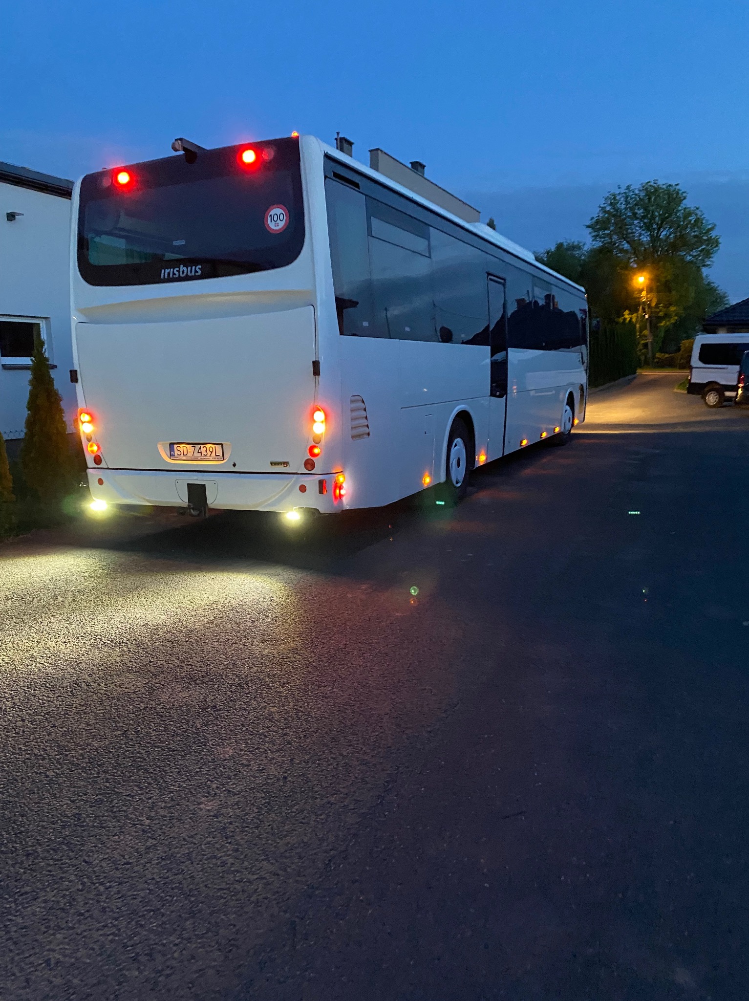 Biały autokar Irisbus, widok z tyłu, oświetlony światłami stopu i cofania, zaparkowany na asfalcie. Widoczny numer rejestracyjny i znak ograniczenia prędkości do 100 km/h.