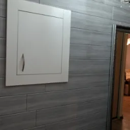 Szara ściana z płytek imitujących drewno, biała klapa rewizyjna z uchwytem, fragment lustra i żyrandola. Nowoczesne wykończenie wnętrza.