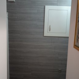 Damian Kontek - Szara ściana wyłożona płytkami imitującymi drewno, z białą szafką rewizyjną i skrzynką elektryczną. Podłoga z paneli drewnopodobnych. Widok z korytarza.