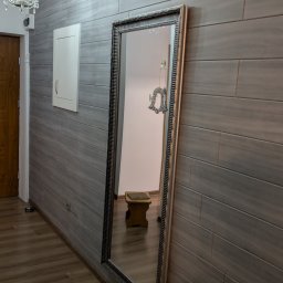 Damian Kontek - Wykończone wnętrze: szara ściana z poziomych paneli, duże lustro w ozdobnej ramie, drewniana podłoga i żyrandol z kryształkami. Widoczne drzwi i szafka.