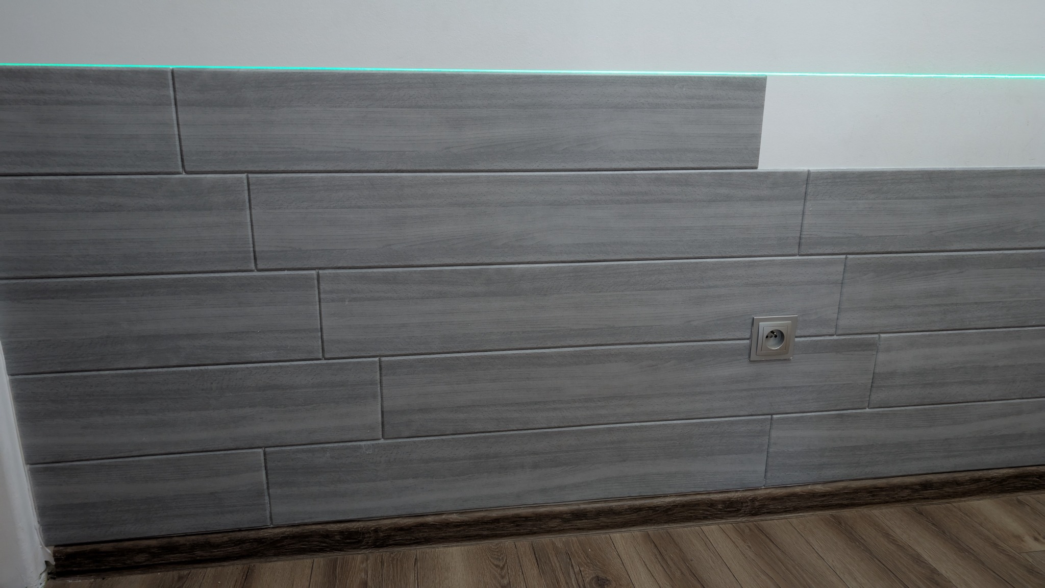 Szare, drewnopodobne płytki ułożone horyzontalnie na ścianie, z widocznym gniazdkiem elektrycznym i zieloną listwą LED. Częściowo widoczna podłoga z paneli.