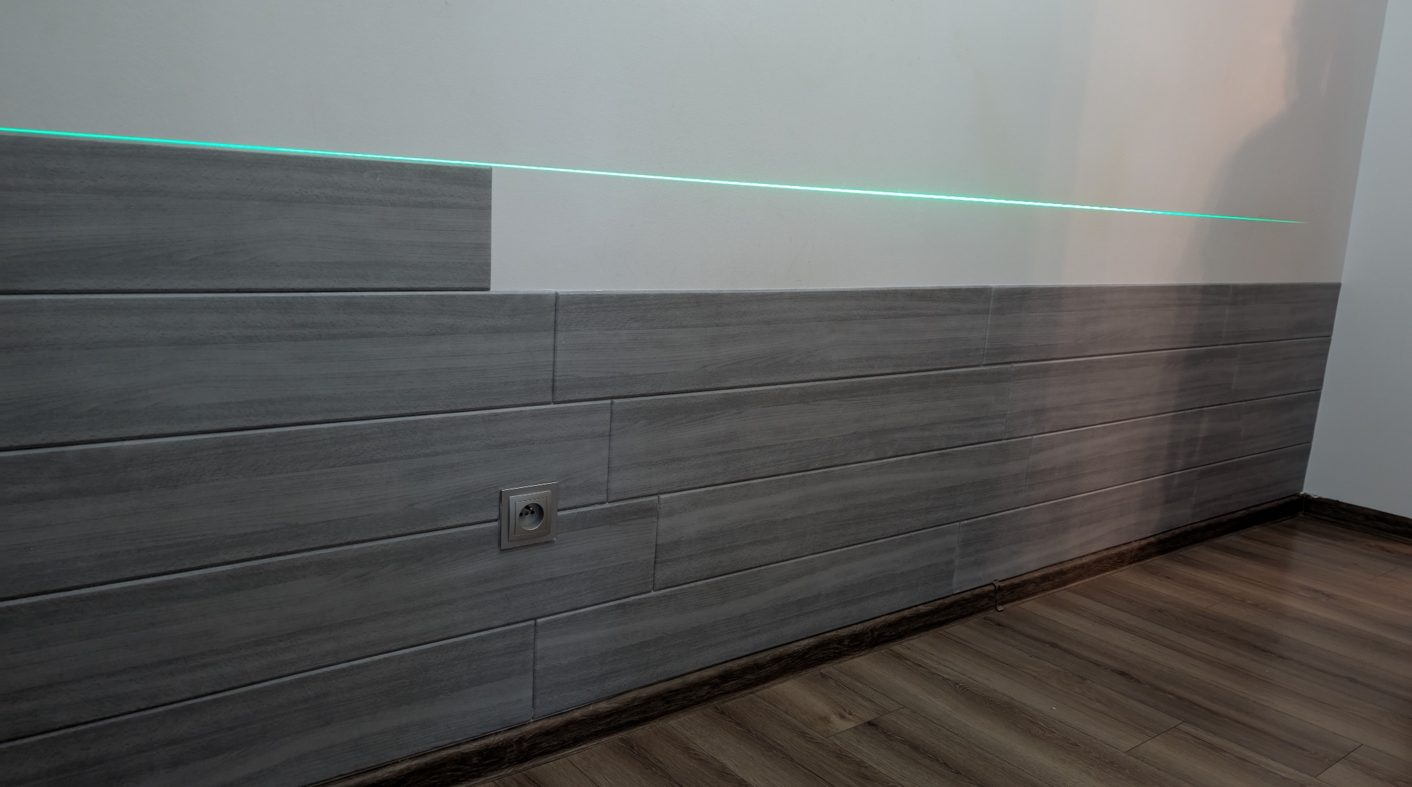 Ściana wyłożona szarymi panelami imitującymi drewno, z widocznym gniazdkiem elektrycznym i poziomą linią lasera, wskazującą na precyzję montażu.