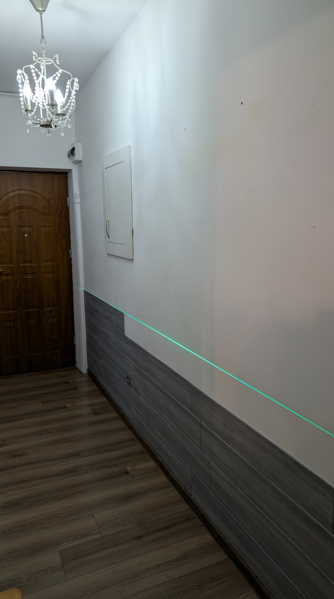 Korytarz po częściowym wyłożeniu ściany szarą płytką imitującą drewno, z widoczną linią lasera wyznaczającą poziom, drewniane drzwi i parkiet.