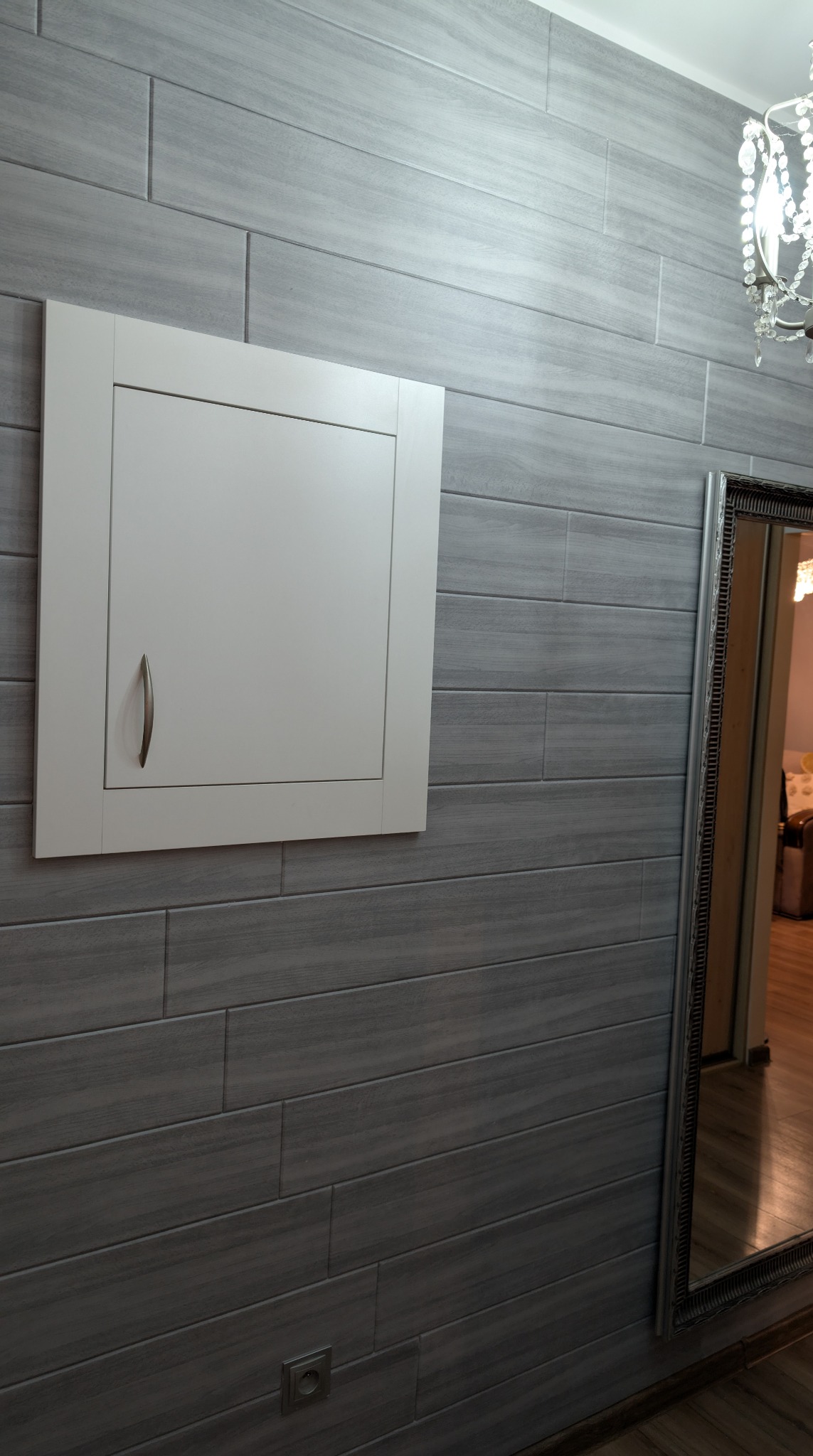Szara ściana z płytek imitujących drewno, biała klapa rewizyjna z uchwytem, fragment lustra i żyrandola. Nowoczesne wykończenie wnętrza.