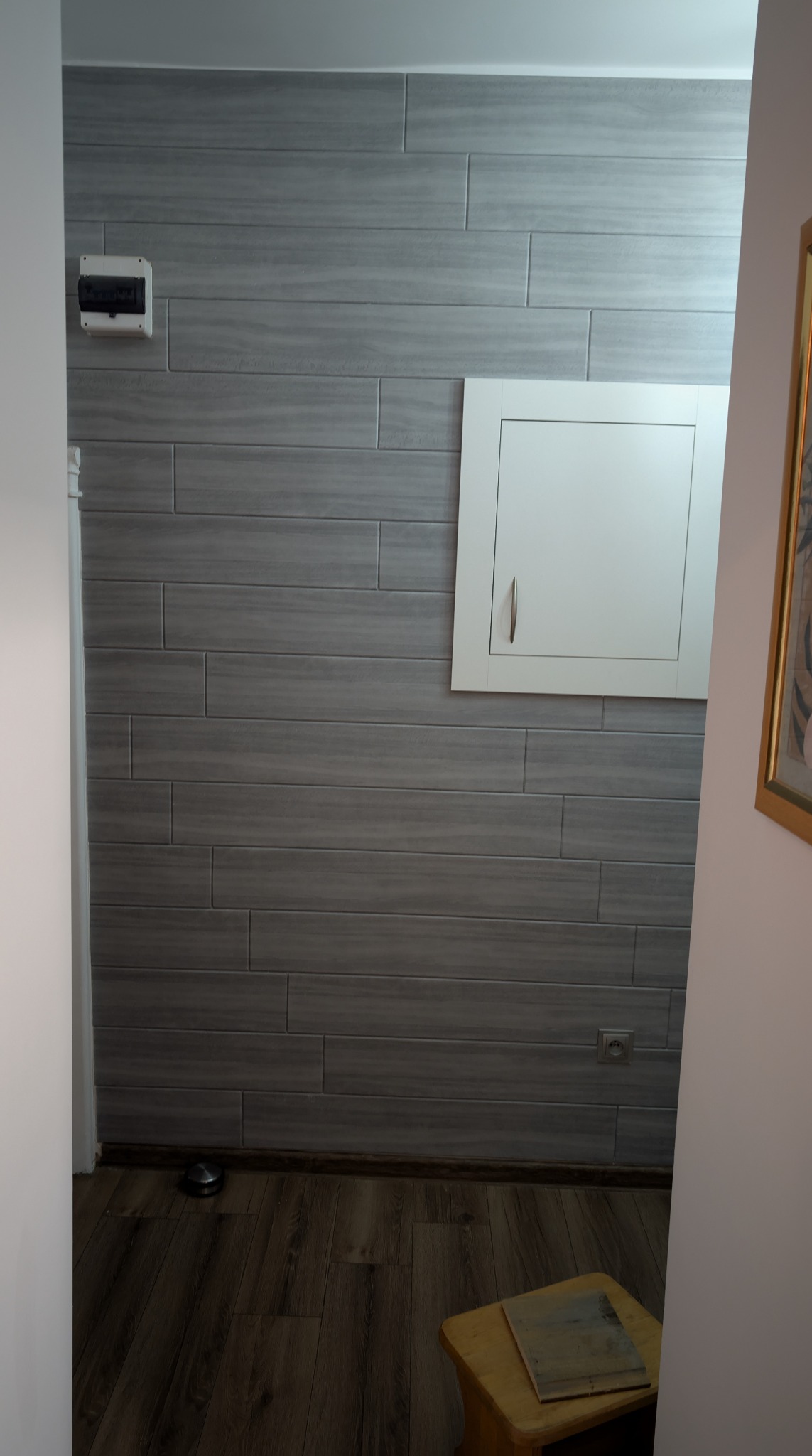 Szara ściana wyłożona płytkami imitującymi drewno, z białą szafką rewizyjną i skrzynką elektryczną. Podłoga z paneli drewnopodobnych. Widok z korytarza.