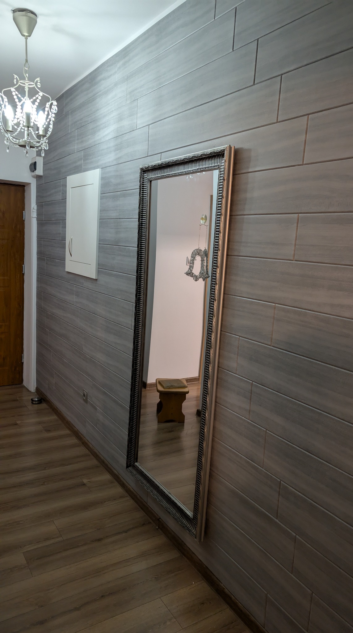 Wykończone wnętrze: szara ściana z poziomych paneli, duże lustro w ozdobnej ramie, drewniana podłoga i żyrandol z kryształkami. Widoczne drzwi i szafka.