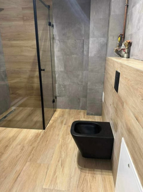 Wykończona łazienka w nowoczesnym stylu: czarna toaleta podwieszana, kabina walk-in, płytki drewnopodobne na podłodze i betonowe na ścianach. Widoczne instalacje.