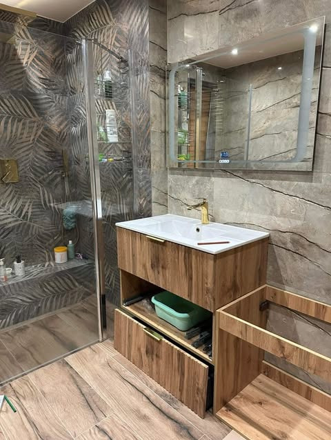 Nowoczesna łazienka z kabiną walk-in, drewnopodobną szafką pod umywalką z otwartą szufladą, złotą armaturą i roślinnym motywem na ścianie.
