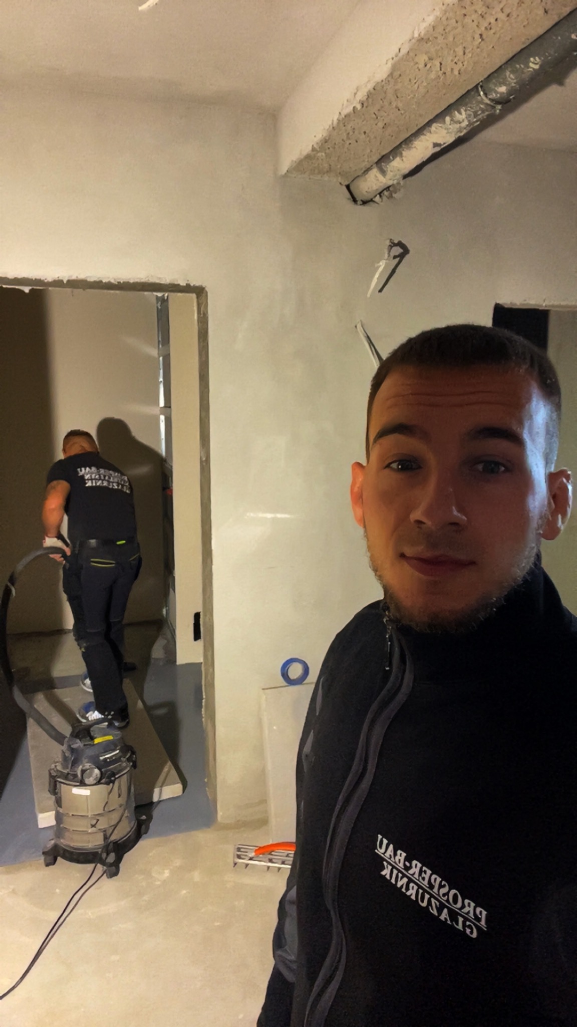 Pracownik sprząta odkurzaczem w remontowanym pomieszczeniu. Na ścianach szara gładź gipsowa. W tle widoczne metalowe profile ścianki działowej.