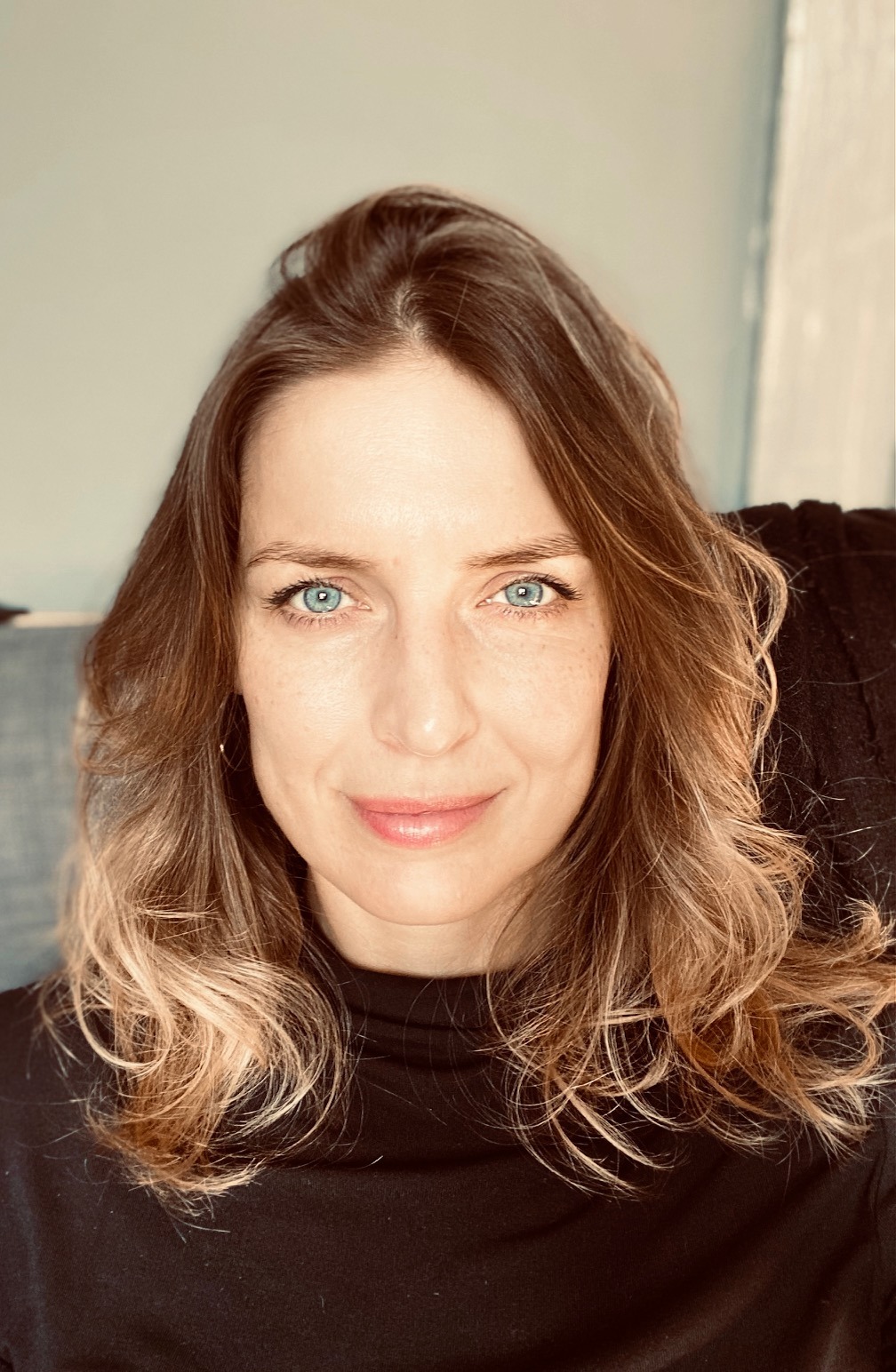 Portret kobiety z długimi, falowanymi włosami w odcieniach blondu i brązu, ubranej w czarny golf, z jasną cerą i niebieskimi oczami, patrzącej wprost.