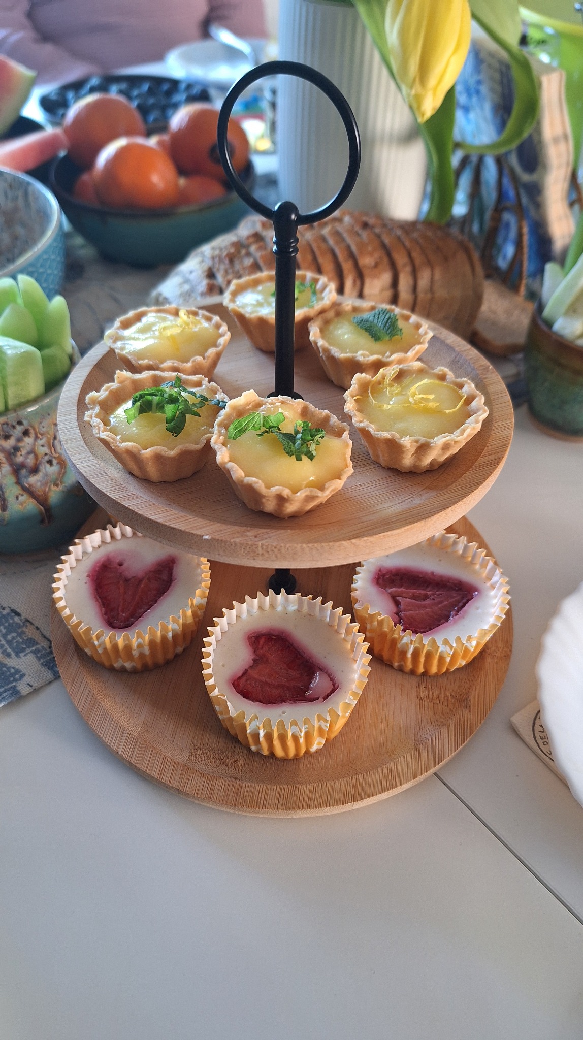 Dwupoziomowa etażerka z babeczkami: cytrynowe z miętą i truskawkowe z serkiem, na stole z owocami i pieczywem. Słodki catering na przyjęcie.