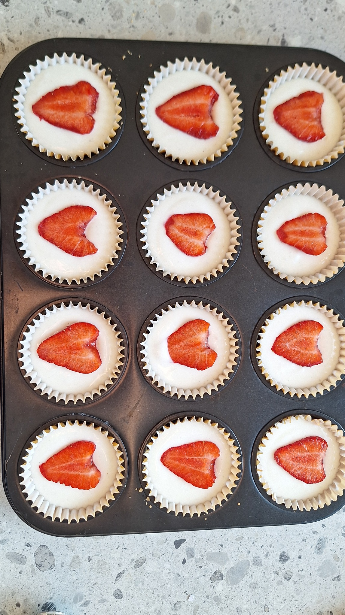 Blacha z babeczkami w papilotkach, wypełnionymi białym kremem i kawałkami truskawek, gotowe do pieczenia. Widok z góry, ostre światło.